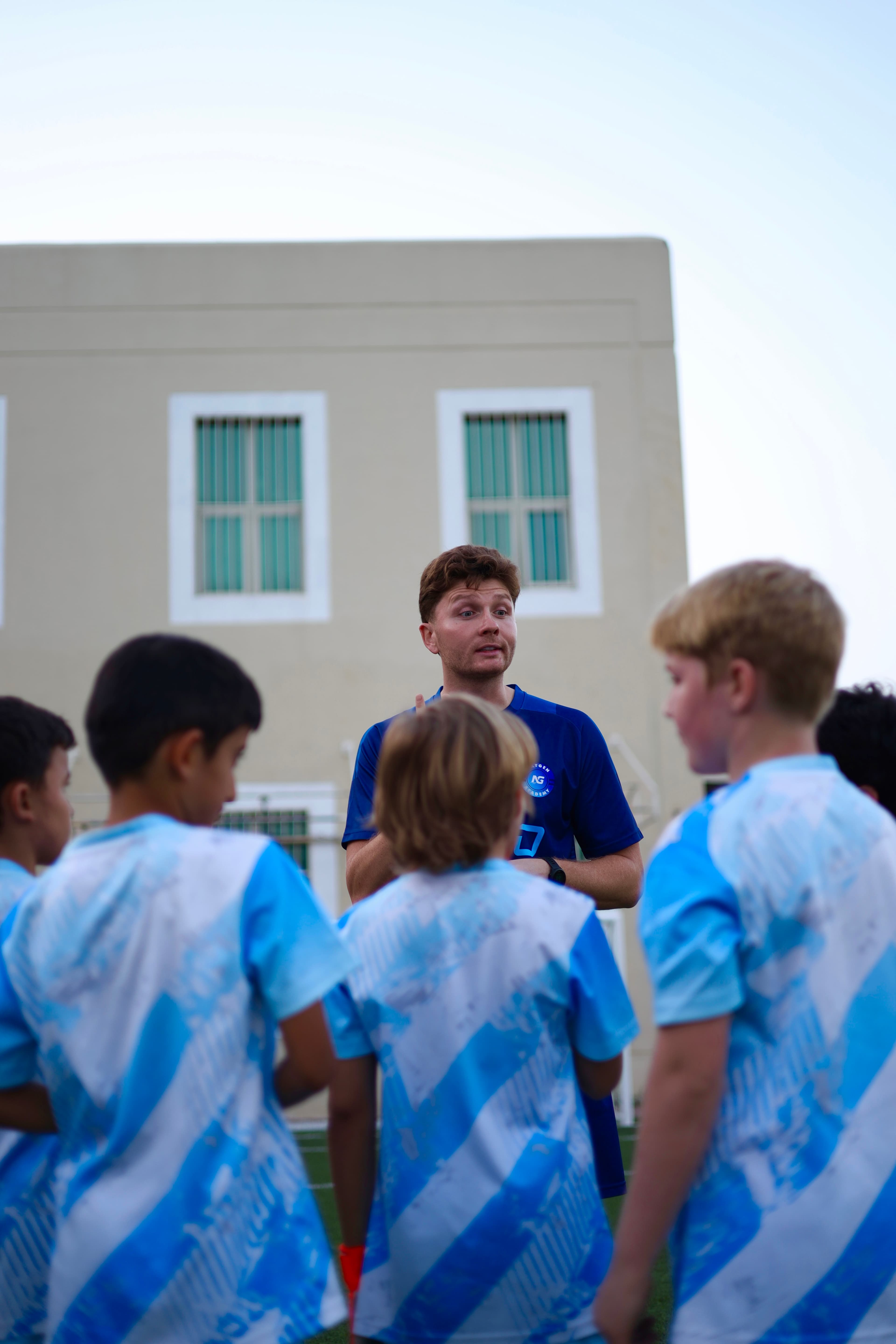 Coach Tommy speaks to NXTGEN players gathered around him on the pitch.